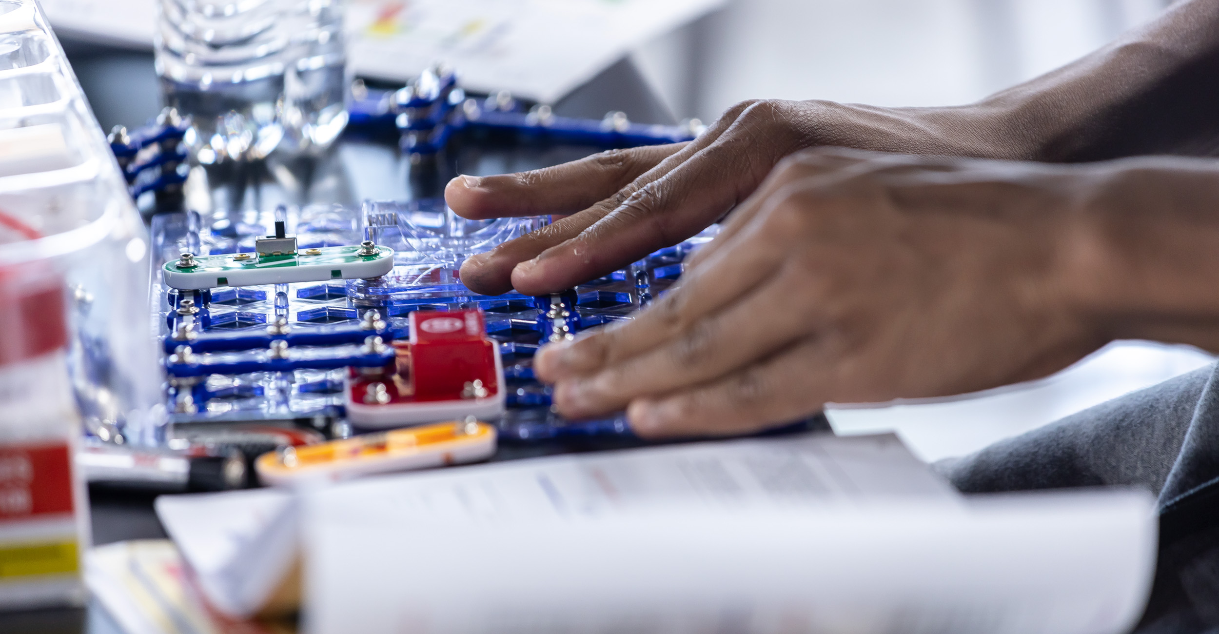 A colorful circuit board with student's hands working to manipulate it into working.