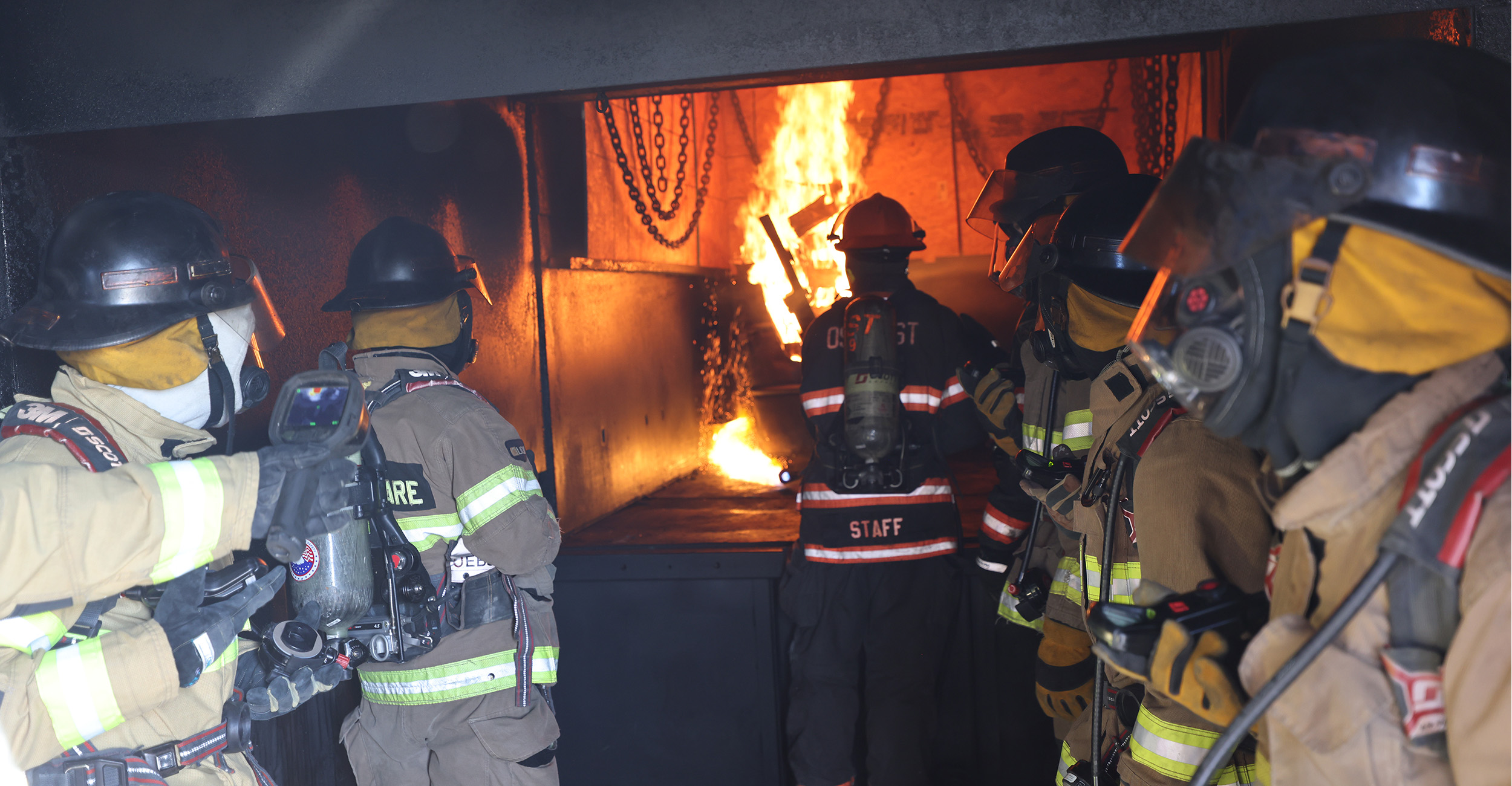 Firefighters sit in a mobile fire lab as part of a training course called Advanced Fire Behavior taught by OSU Fire Service Training. One is lighting a bundle of wood that starts to burn, while the rest of the firefighters prepare to experience extreme heat inside the lab. This training allows them to feel how hot certain temperatures are in a controlled environment.