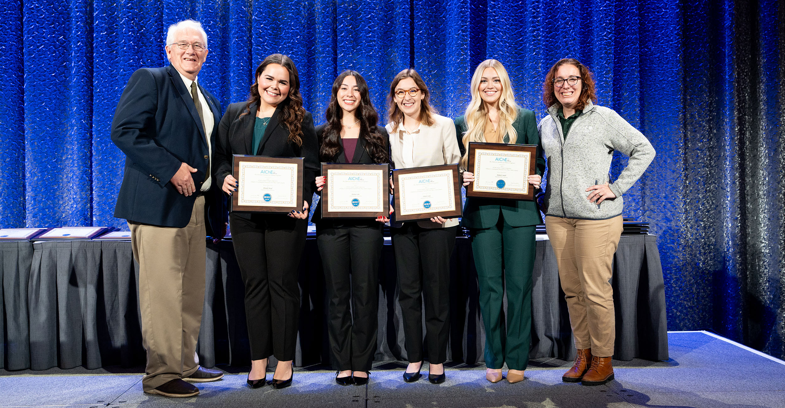 Brandi Head, Kenna Lam, Nikole Salas and Blakely Smith stand with representatives from AIChE holding framed plaques on a stage while all dressed in fancy clothing.