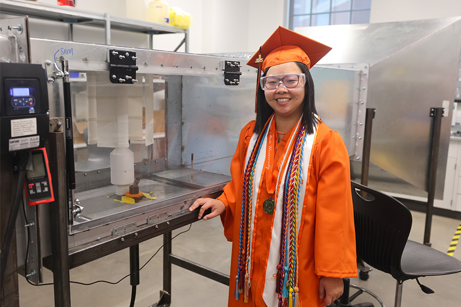 A student in graduation regalia stands for a portrait in a lab setting.