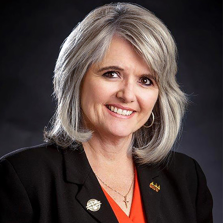 A woman with shoulder-length gray hair wearing a black blazer, an orange top and a small cross necklace.