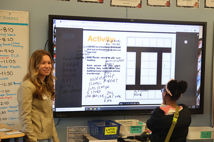 Students in the process of teaching students smile at the front of a classroom. 