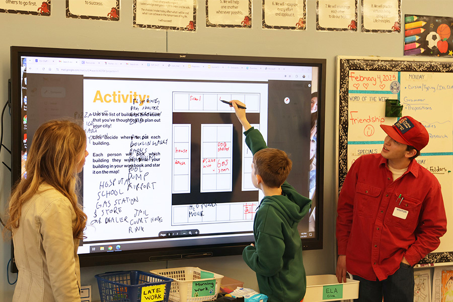 A student writes on the board as part of an assignment while two older students look on. 