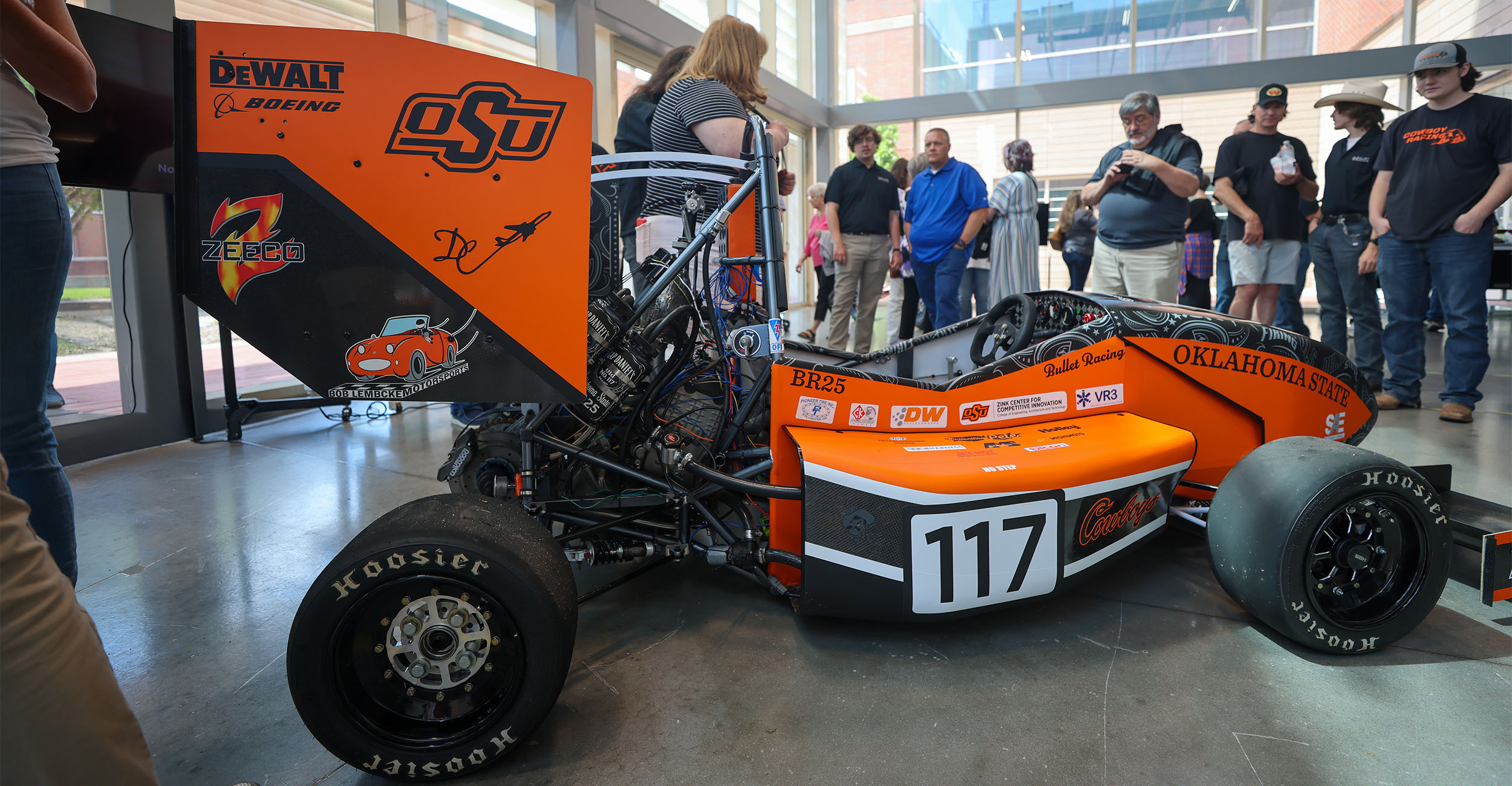 An orange and black formula race car on display in a modern laboratory, with individuals in the background looking at the vehicle.
