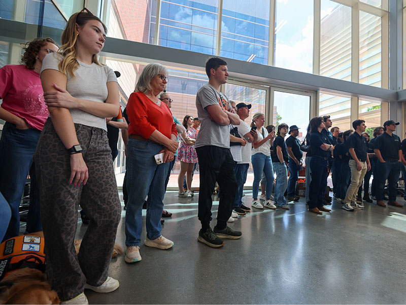 A large crowd gathered in a modern laboratory looking towards a veiled vehicle.