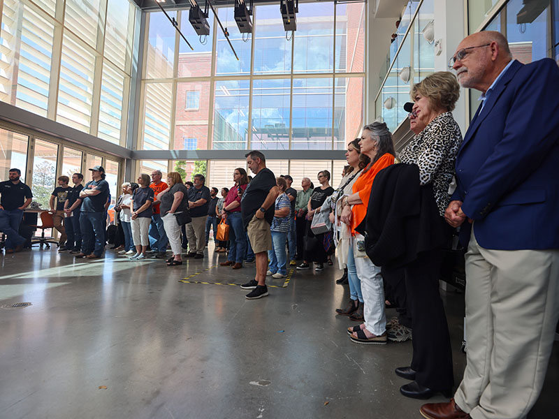 A large crowd gathered in a modern laboratory looking towards a veiled vehicle.