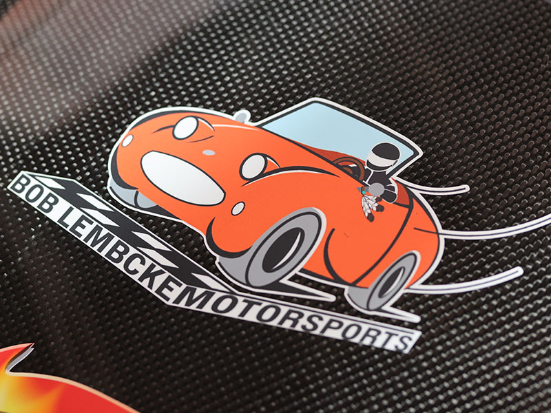 A decal of an orange car with Bob Lembcke Motorsports under it and three feathers hanging from the mirror.