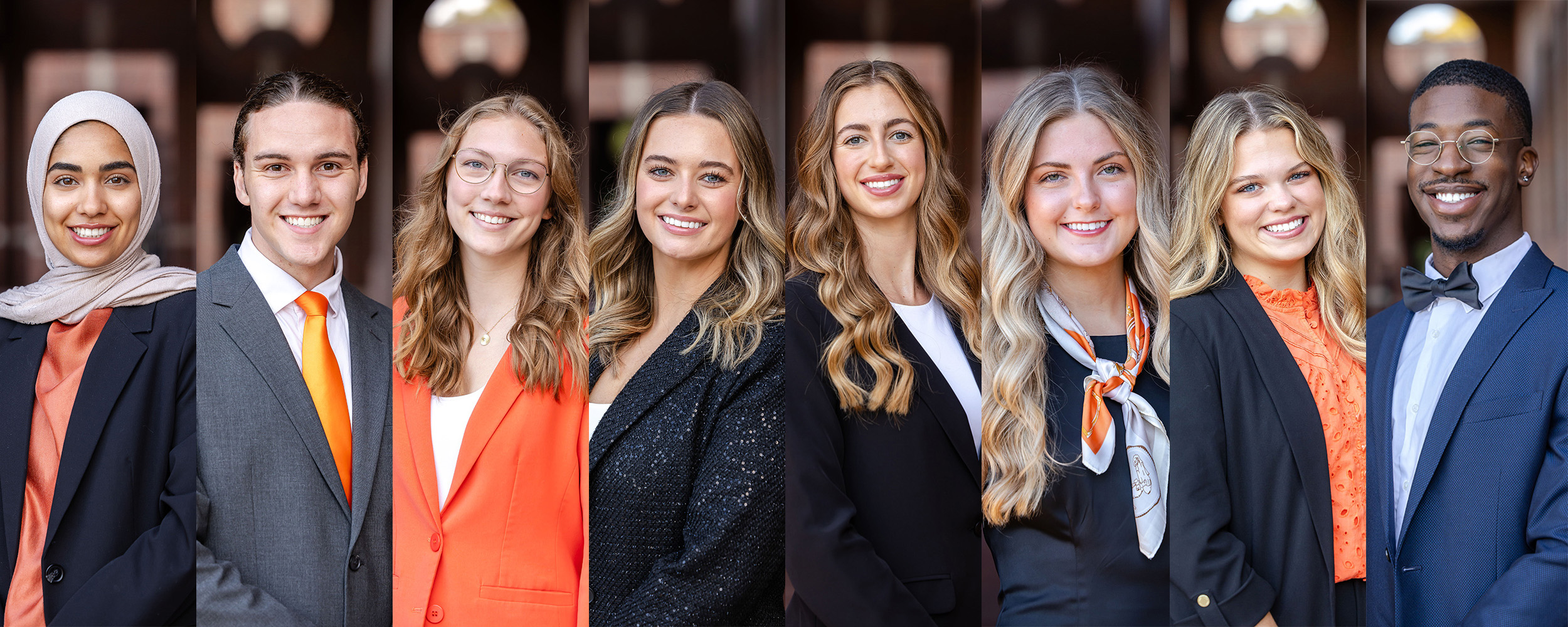 Group of eight people standing side by side in formal and business attire, wearing black, gray, and orange clothing with OSU-themed accents such as scarves and ties. They are posed outdoors in front of a building with round hanging lights.