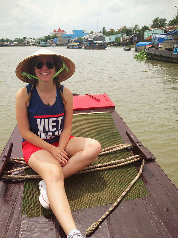 A smiling individual sits in a canoe with a large brim hat on and a tank top that says Vietnam.