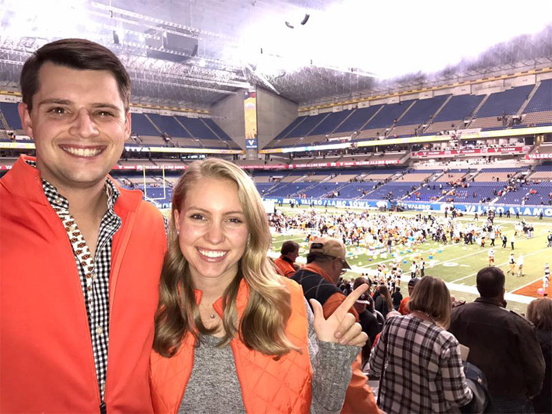 Two people dressed in OSU colored clothing pose at a football game from seats near the field.