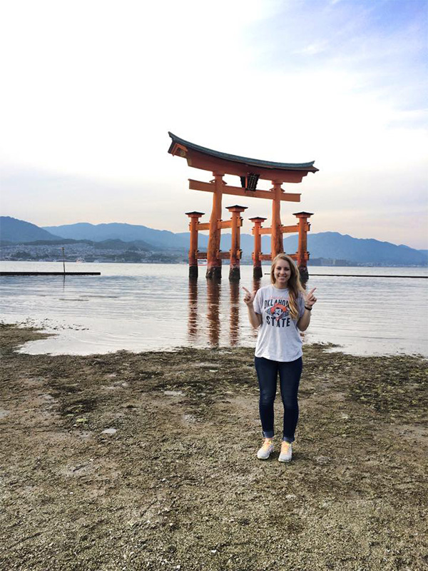 A young college student standing on a shore in Japan with the floating torii gate of Itsukushima Shrine in the background.