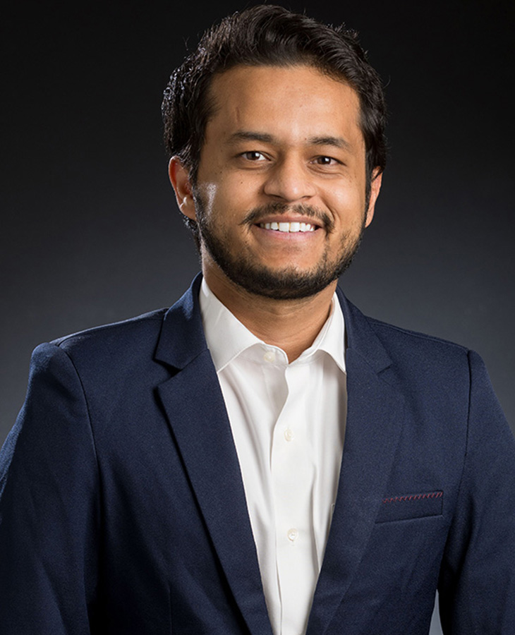 A professional headshot of a man dressed in a navy blue suit with a black background.
