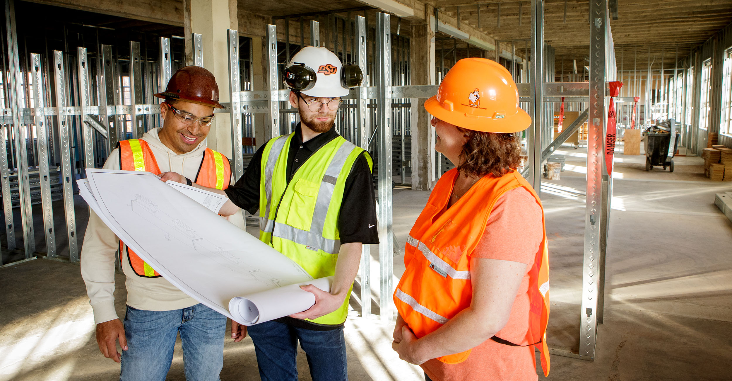 Three individuals wearing reflective vests and hard hats look over blueprints in a construction area.