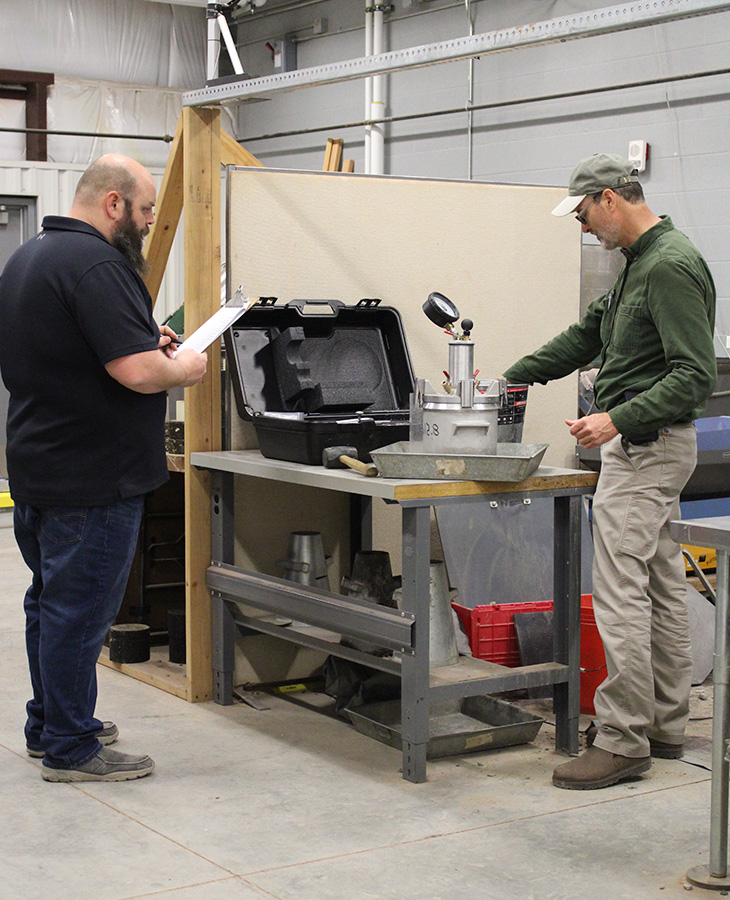 Two individuals in a workshop setting, with one person holding documents and standing beside a toolbox, while the other interacts with equipment on a workbench.