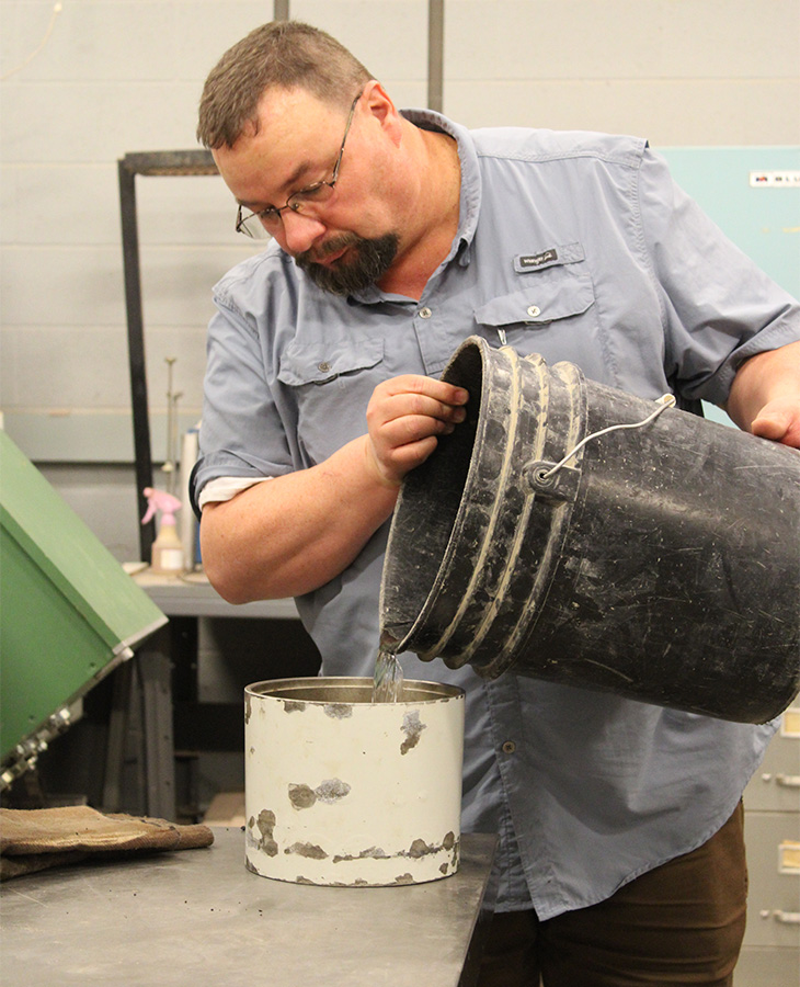 A person wearing a blue shirt and glasses pouring contents from a black bucket into a white container in a workshop setting.