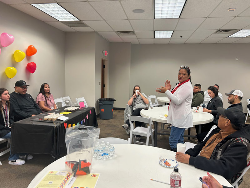 A person stands speaking to a group seated around tables at an indoor gathering with colorful balloons and refreshments.