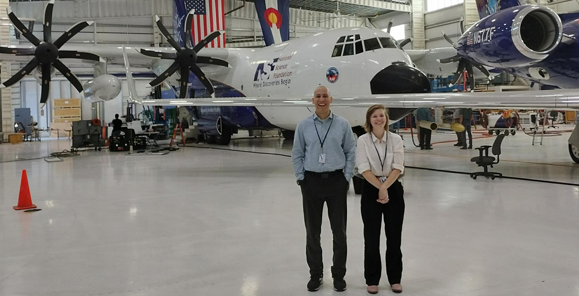 Two professionally dressed people stand in an aircraft hanger with two large aircraft in the background.