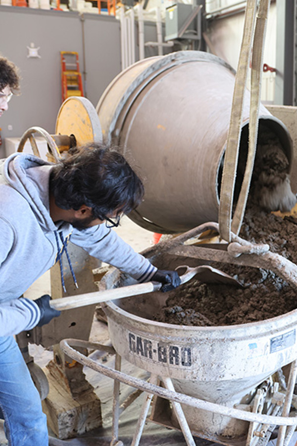 A man wearing a hoodie uses a shovel to move around concrete in a metal cylinder.