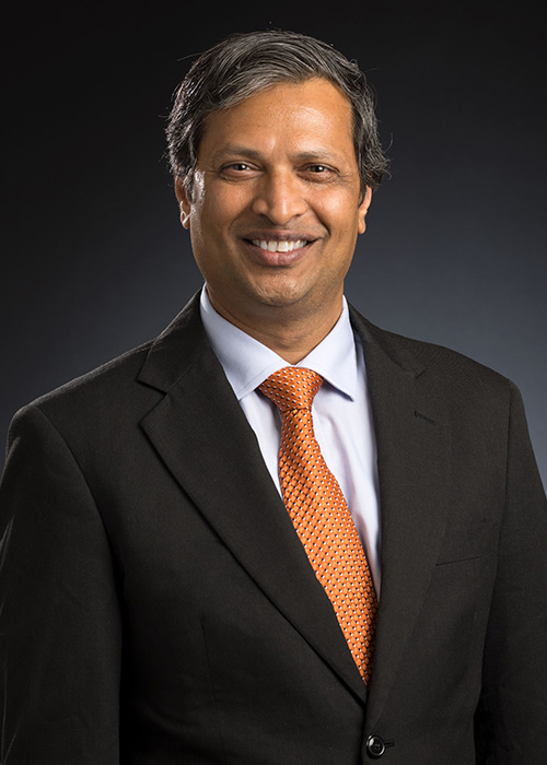 A professional headshot of a man in a navy blue suit and orange tie.