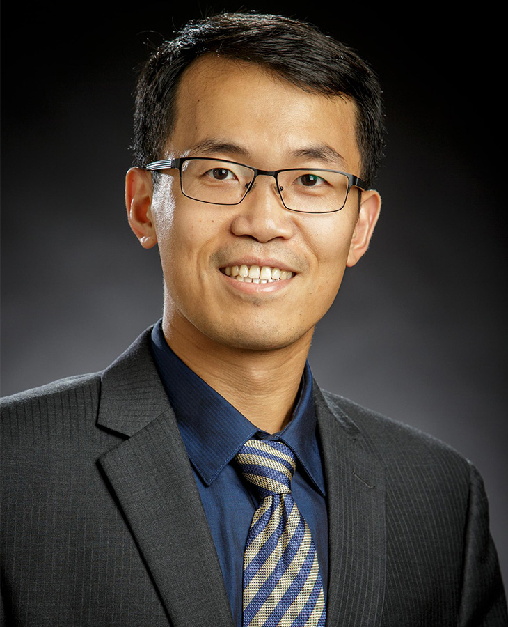 A professional headshot of a man smiling wearing a suit and tie with a black background.
