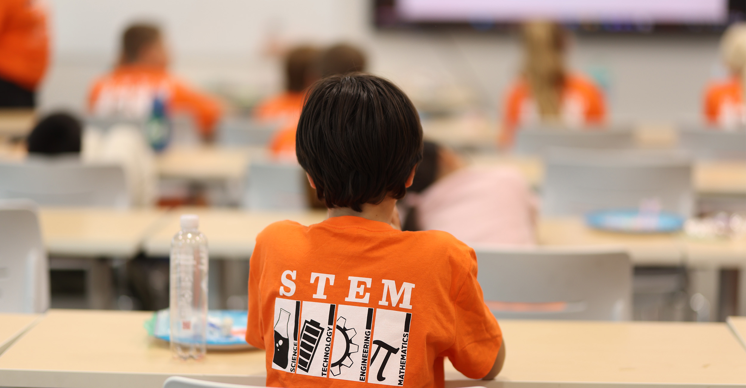 A child wearing an orange shirt with the word 'STEM' and science icons on the back sits at a desk in a classroom. Other children in matching shirts are seated at desks facing a large screen at the front. A water bottle and plate are on the child's desk.