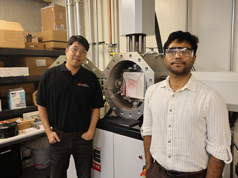 Dr. Hunjoo Lee, a professor in the Oklahoma State University School of Chemical Engineering, and his student Vishesh Bhadariya stand beside a large testing machine in Lee’s lab. The equipment appears to be used for material or fracture testing, and shelves with research supplies and boxes are visible in the background.