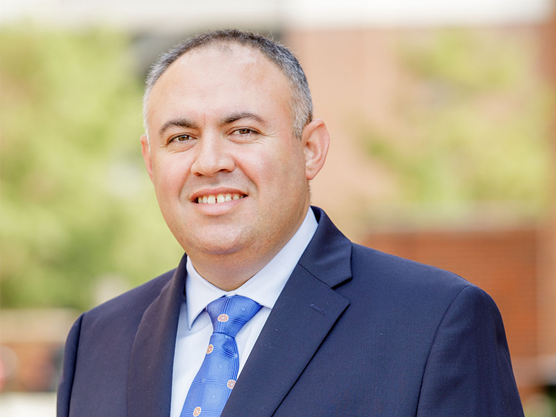 A man dressed in a navy blue suit poses for a professional headshot with an outdoor background.