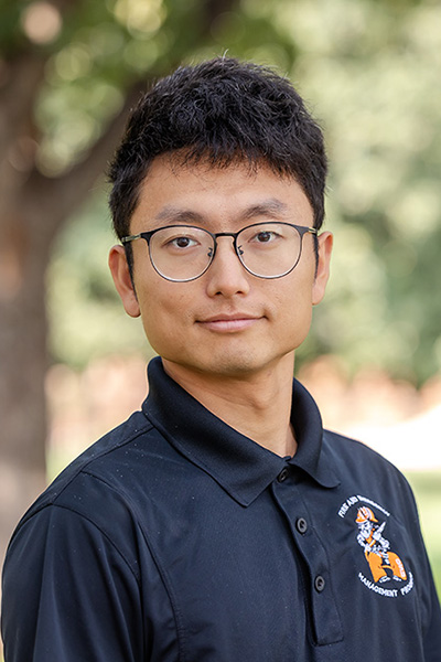 Dr. Chen Chen of the OSU School of Fire, Construction and Emergency Management is photographed in a polo with the OSU logo outside for a staff portrait.