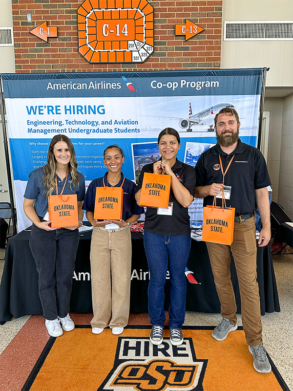 Four people holding orange Oklahoma State bags stand at a career fair in front of an American Airlines booth.