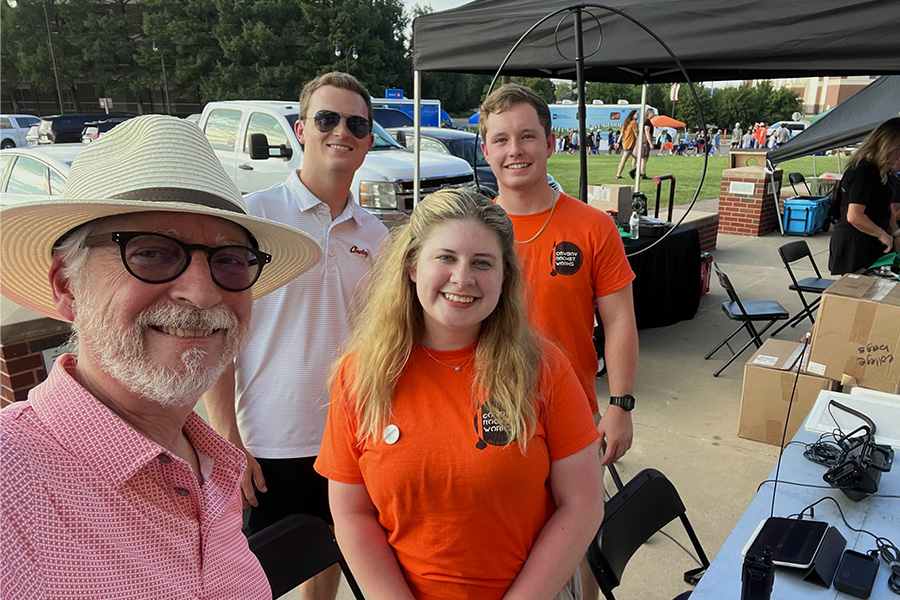 Four people pose for a group photo while standing outdoors near a table with radio equipment in use. 