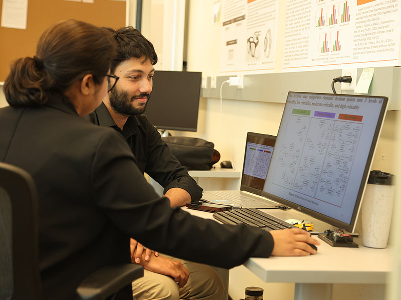 A student smiles as he looks over research on a computer screen with his professor.