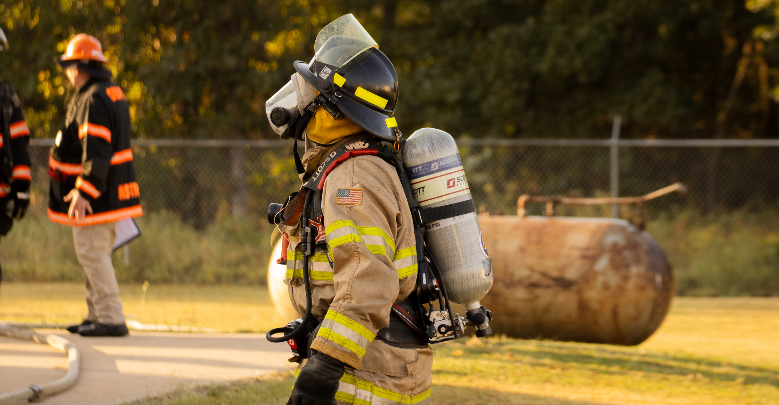 A firefighter in full gear stands outside a burning building. You can see other firemen in the background in OSU FST training gear.