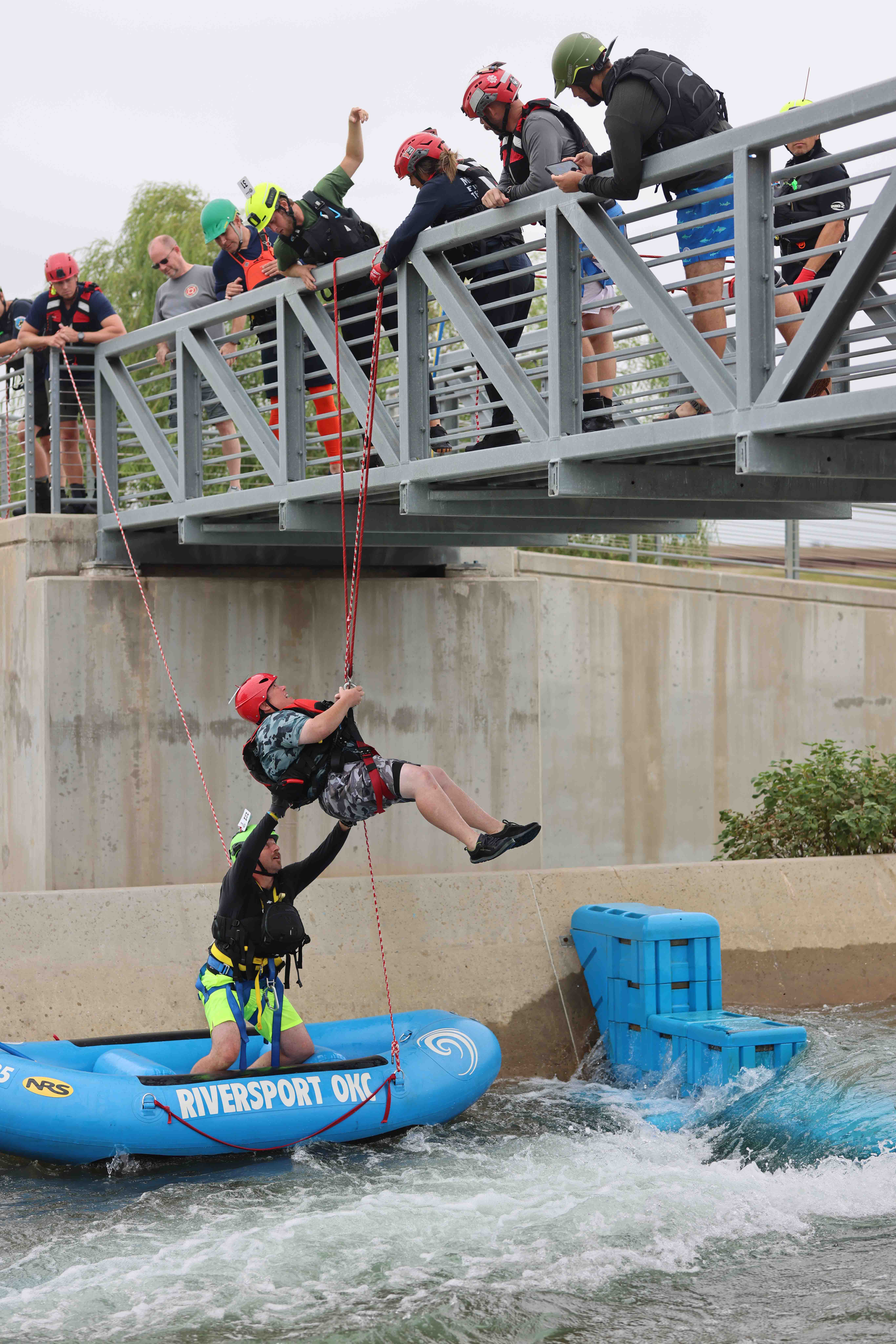 A man standing in a blue raft that is in a river with rapids assists a person in a harness that is being pulled up to the bride overhead. Several first responders stand on the bridge waiting.