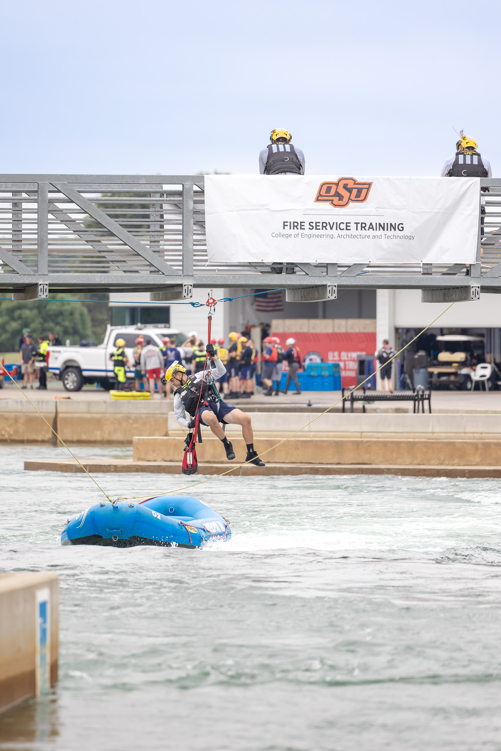 A first responder, held by a harness, hangs over a blue raft in a river.