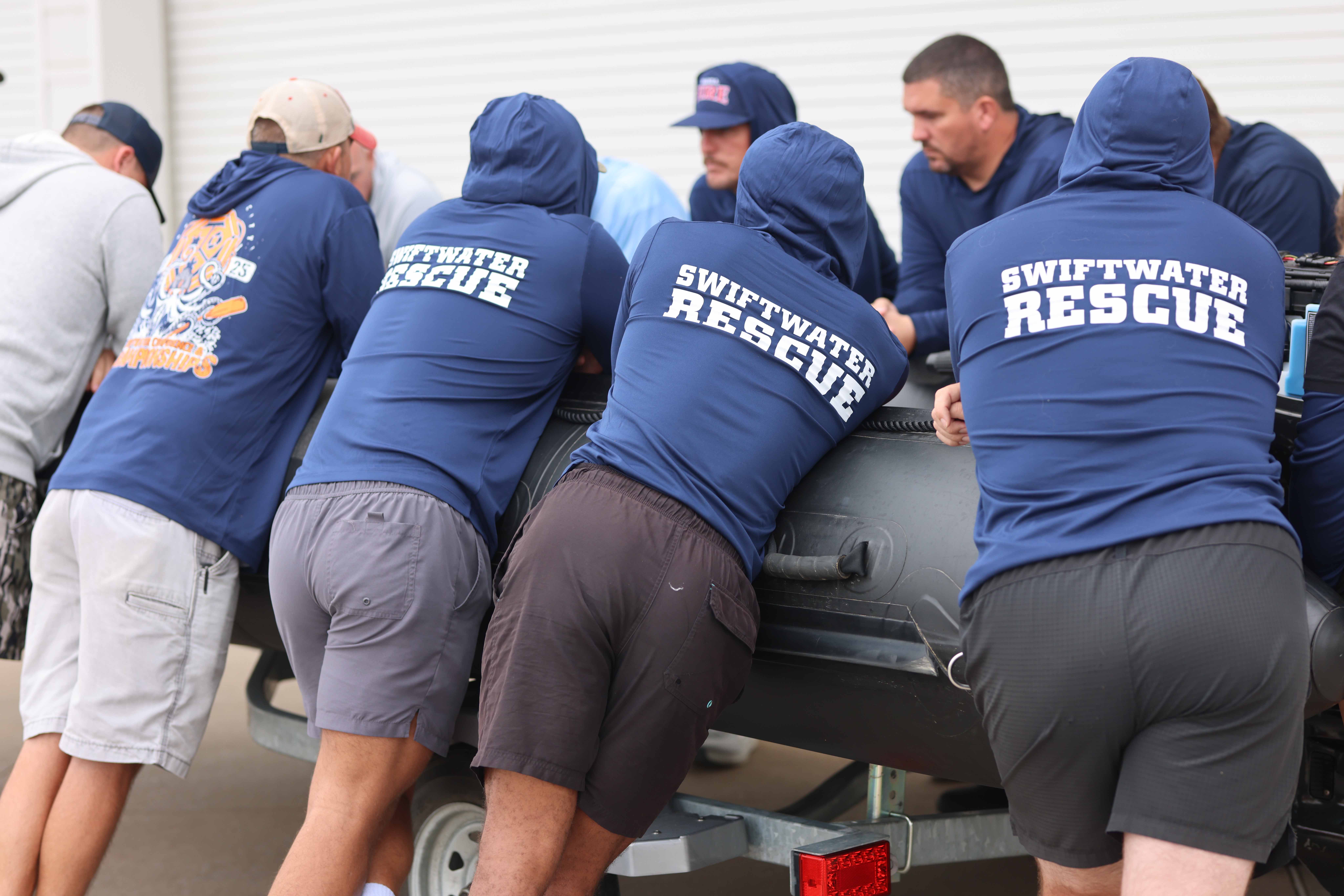 A group of people wearing navy hoodies that say swiftwater rescue on the back gather around a black raft.