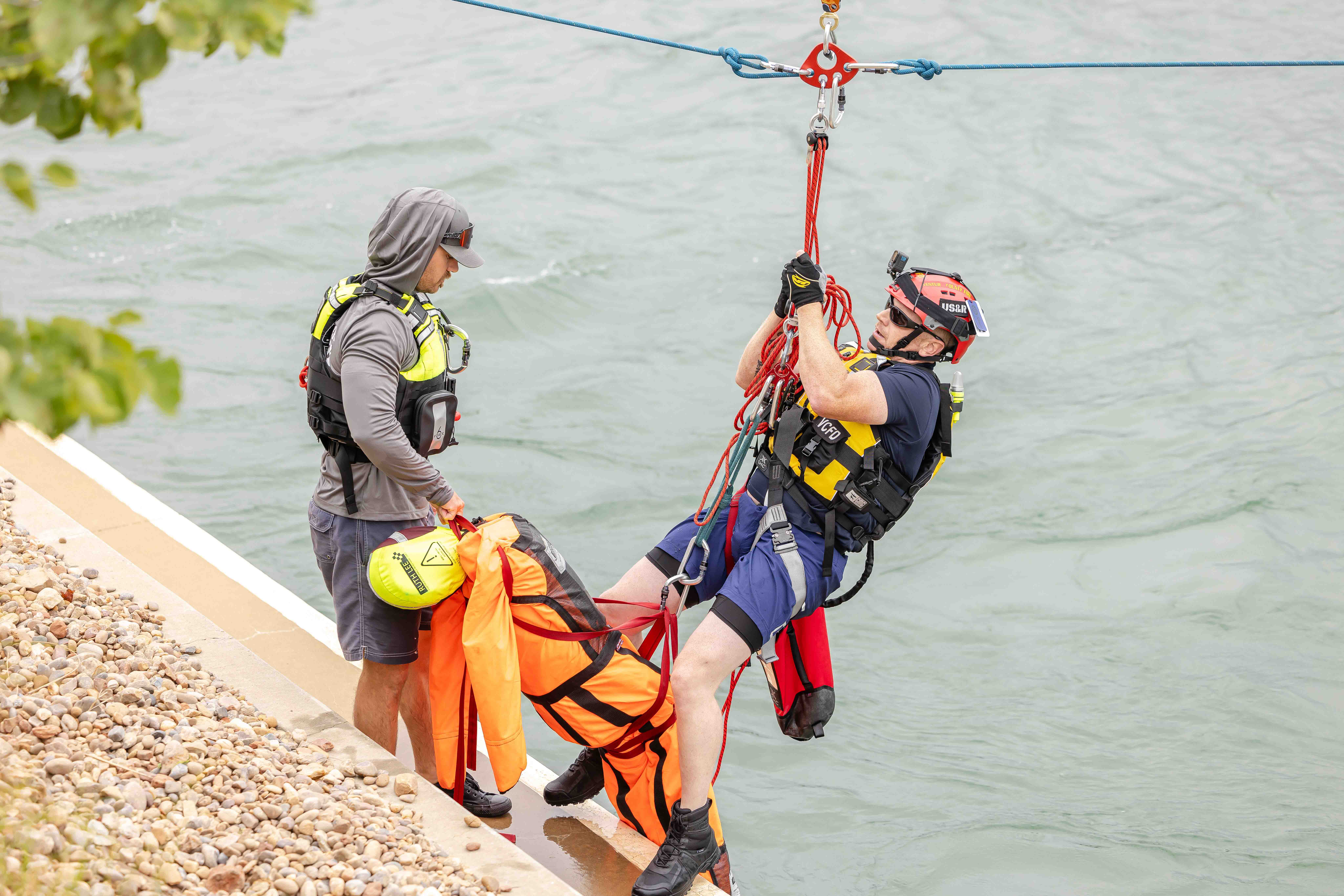 Two men in life vests and helmets are suspended over a river while holding a human dummy.