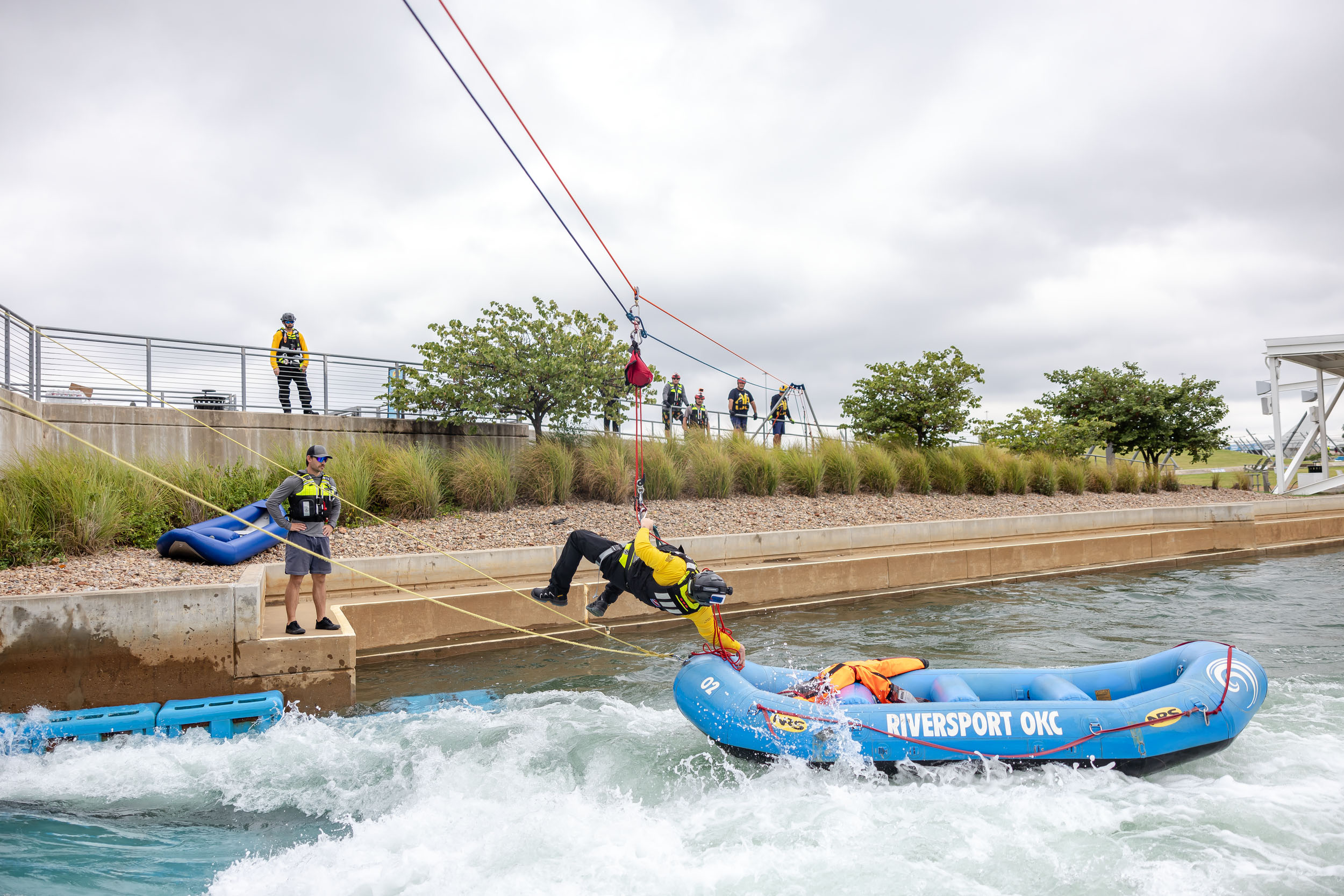 A first responder hangs from a harness while reaching for a dummie in a blue raft that is moving in rapids.
