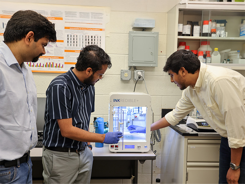 Three OSU researchers stand around a printing testbed to inspect a sample.
