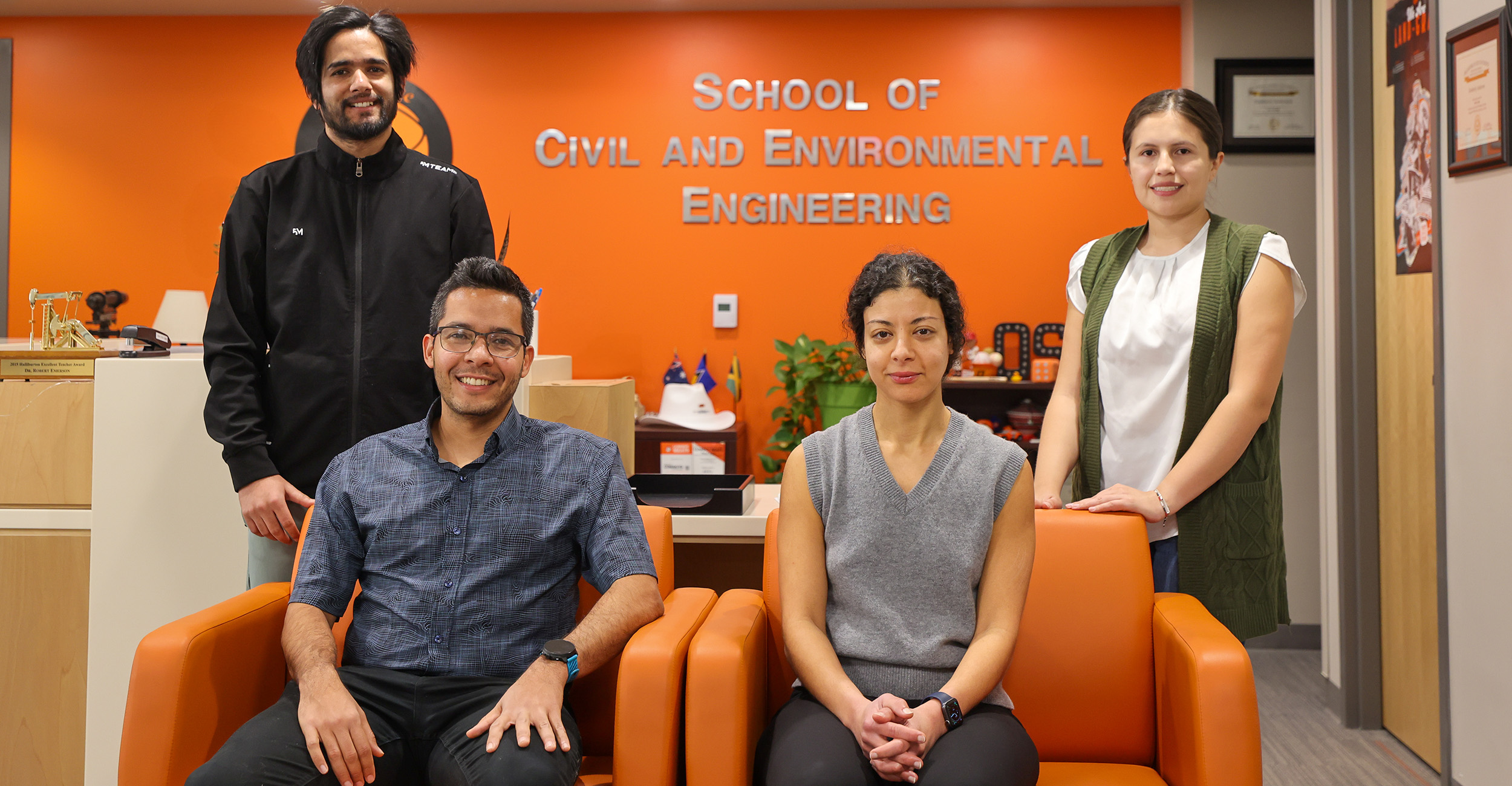 Four people pose in front of a School of Civil and Environmental Engineering, two sit in orange chairs and two stand behind the chairs.