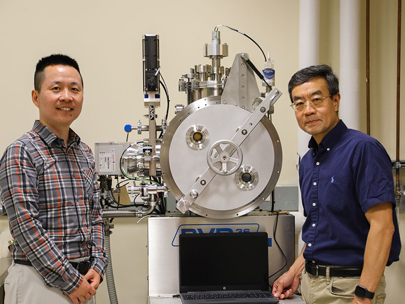 Two men dressed in business casual clothing stand next to a piece of scientific equipment in a lab.