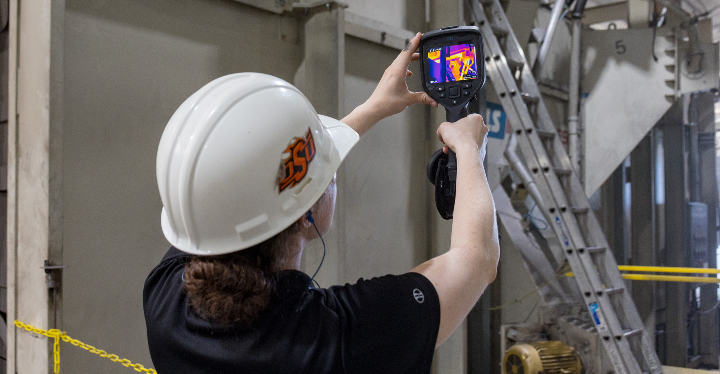 An individual holds up a power consumption meter and the screen displays a thermal imaging display.