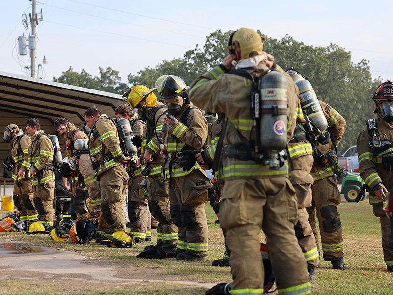 A group of firefighter dressed in full gear stand together looking exhausted. Some have removed their helmets and most are looking towards the ground. 