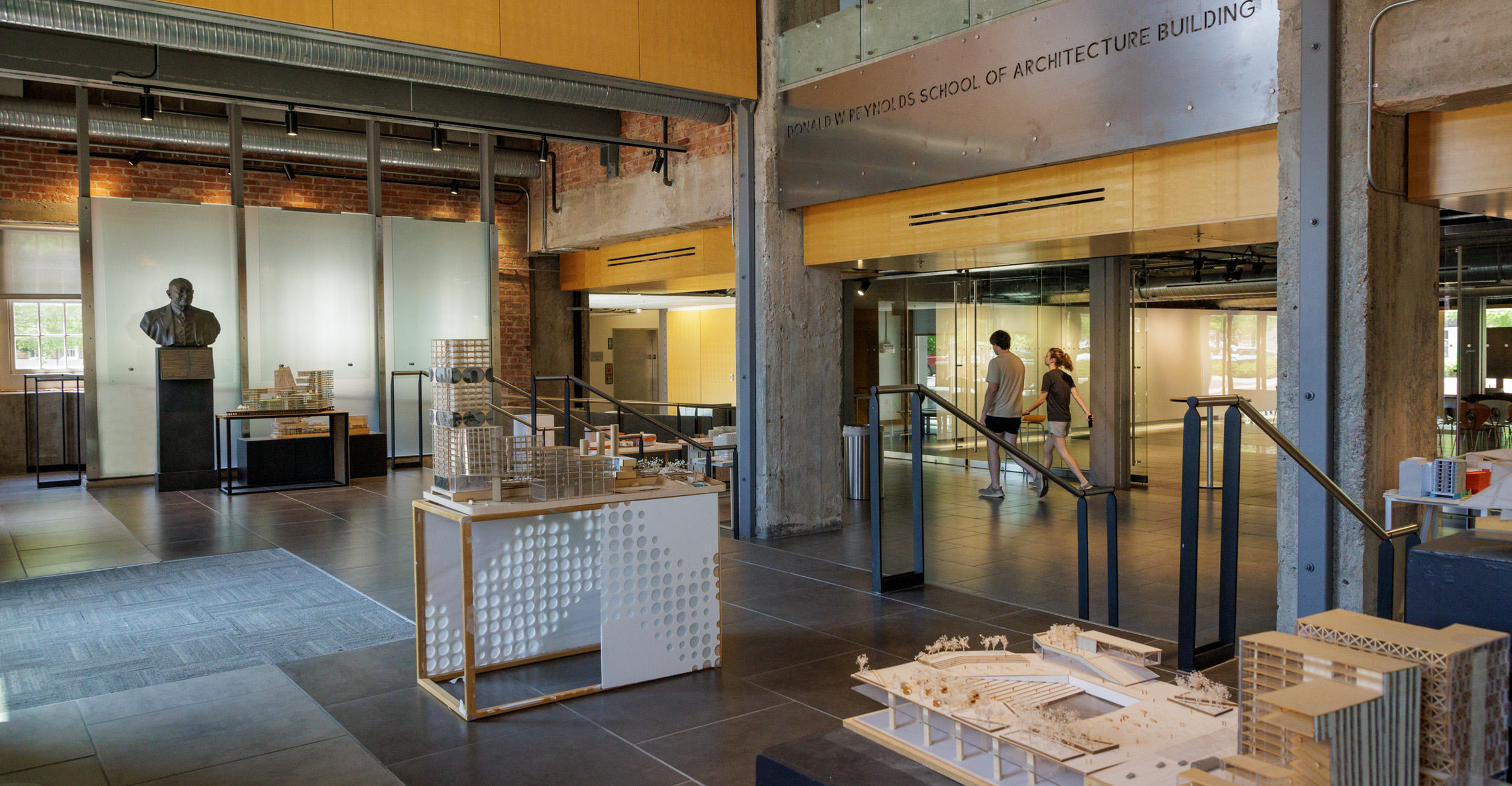 A photo of the front lobby entrance to the School of Architecture. Maquettes can be seen in the foreground, a large metal sign with the school's name can be seen in addition to students walking past glass doors in the background.