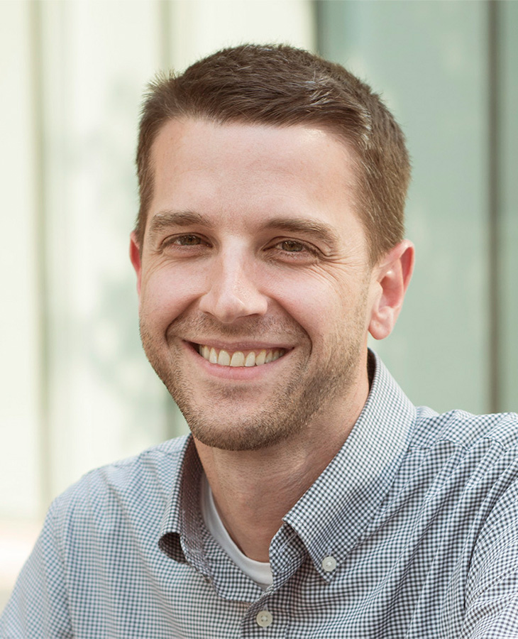 A professional headshot of a man wearing a blue checkered button up shirt.