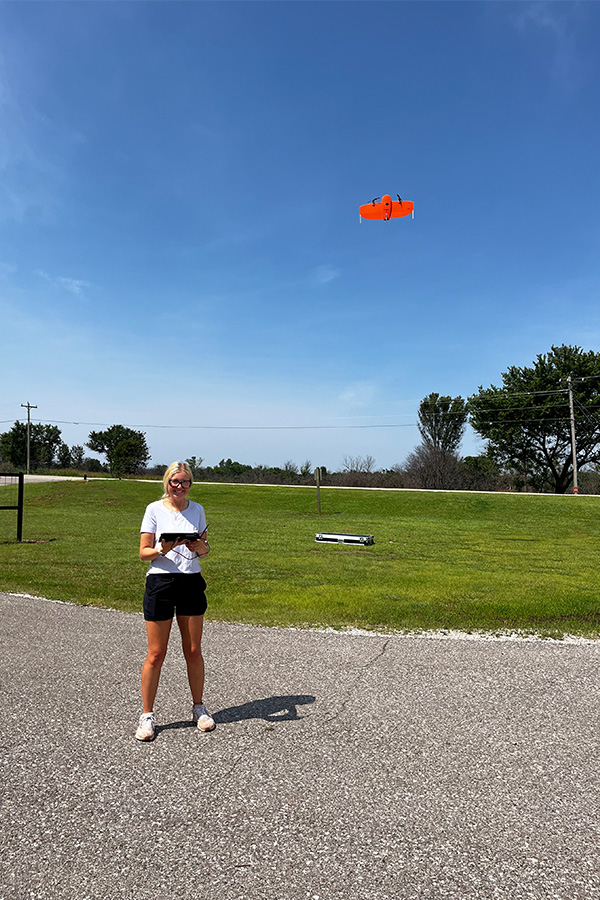 A woman in a parking lot enjoys flying a drone, which hovers in the air above her, set against a sunny backdrop.