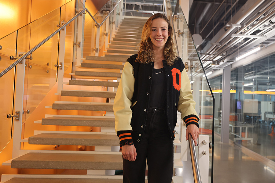 A woman wearing a letterman jacket poses for a photo on a staircase.