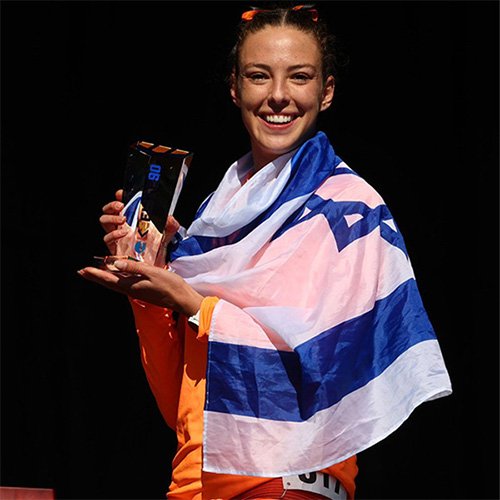 A woman stands holding a trophy from a track competition while an Israel flag is around her shoulders.