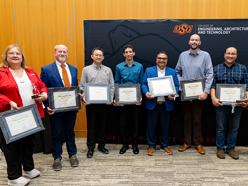A line of individuals stand in front of an Oklahoma State University College of Engineering, Architecture and Technology backdrop, holding framed certificates during a formal recognition or service award presentation.