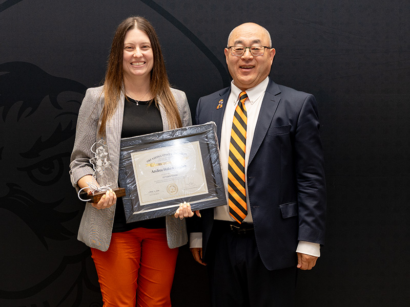 Two individuals stand in front of an Oklahoma State University College of Engineering, Architecture and Technology backdrop, with one holding a framed certificate and a small trophy during a formal recognition or service award presentation.