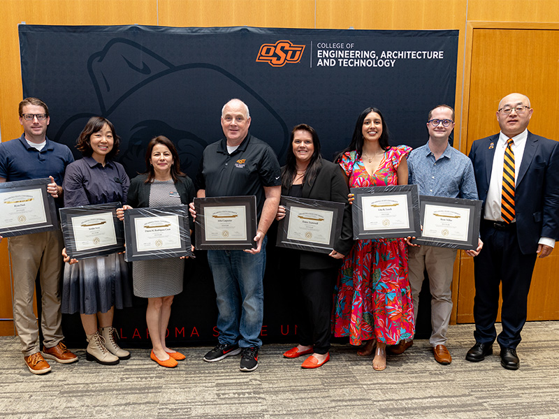 A group of individuals stand in front of an Oklahoma State University College of Engineering, Architecture and Technology backdrop, each holding a framed certificate, indicating a formal recognition or award presentation.